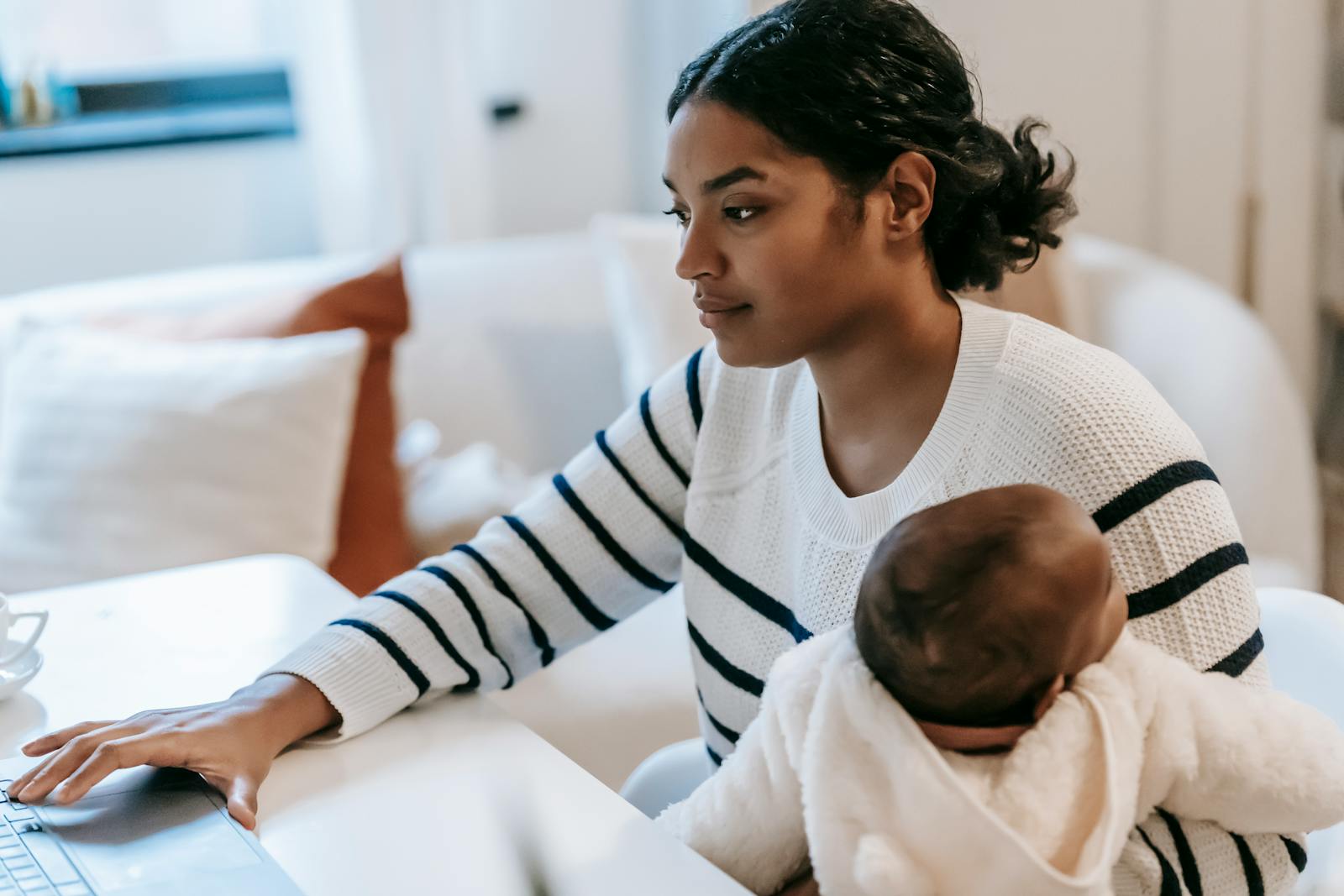 A Black mother holding her baby on her lap while working on her laptop at home